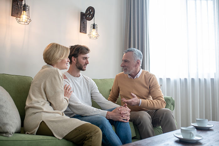 A groom, his mother, and father sit on a green couch, discussing a wedding and the groom's bride's gay brother.