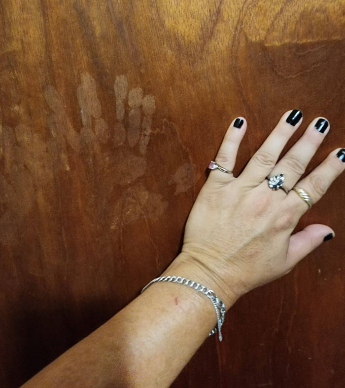 A person's hand with black nail polish next to creepy and confusing handprints on a wooden door, needing a priest.
