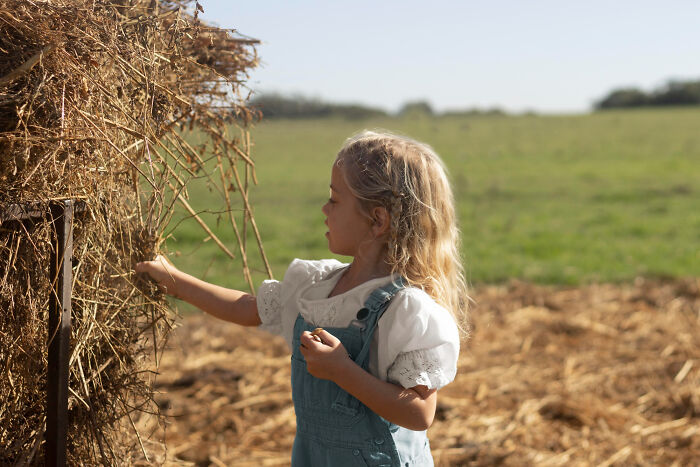 A young girl with blonde hair in overalls interacts with a haystack, enjoying childhood experiences.