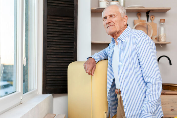 Elderly man in a striped shirt leaning on a yellow refrigerator, reflecting on childhood experiences.