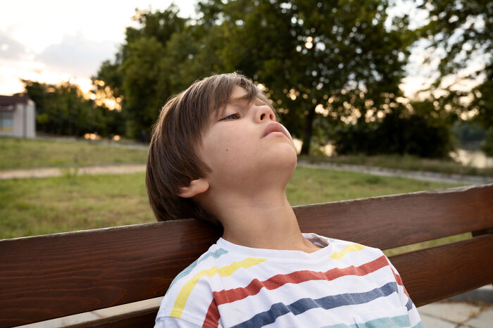 A young boy leans back on a park bench, looking upwards, deep in thought about childhood experiences.