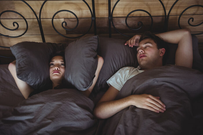 A woman covers her ears with pillows while a man sleeps soundly next to her, depicting a relatable childhood experience.