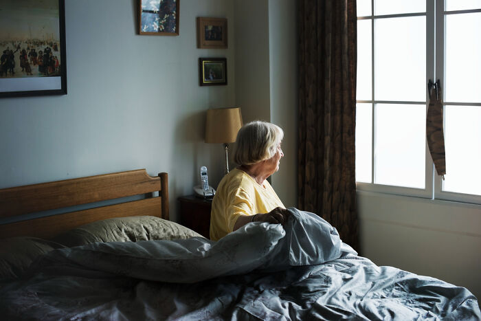 Elderly woman sitting on a bed looking out a window, remembering childhood experiences.