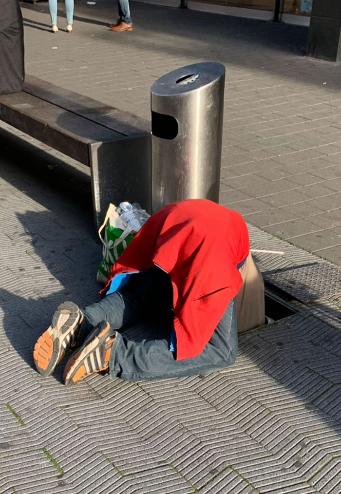 A person in a red jacket and jeans with their head inside a storm drain, next to a trash can, an unusual camera roll photo.