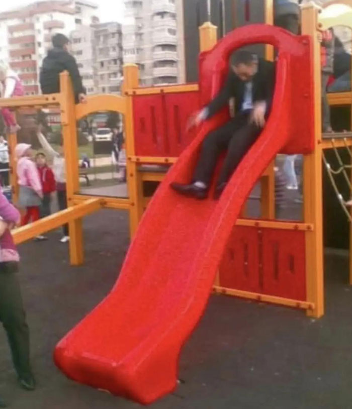 Camera roll photos capture a man in a suit sliding down a red playground slide, an unexpected scene in an outdoor play area.