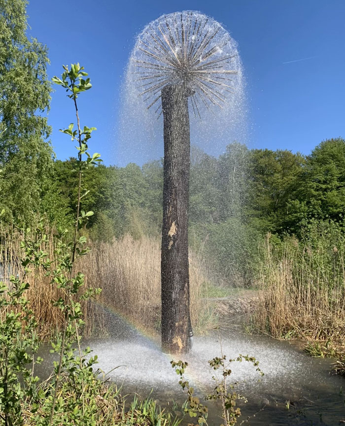 A camera roll photo of a unique tree-like fountain spraying water in a sunny outdoor setting with a rainbow.