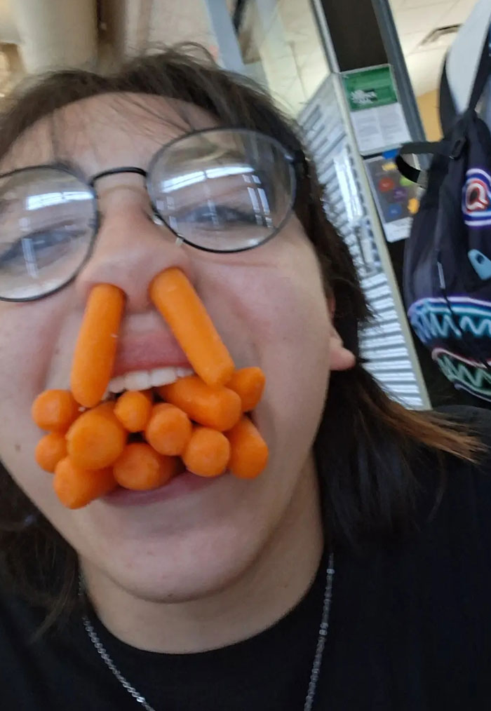 A person with glasses, with baby carrots stuck in their nose and mouth, a weird camera roll photo needing explanation.