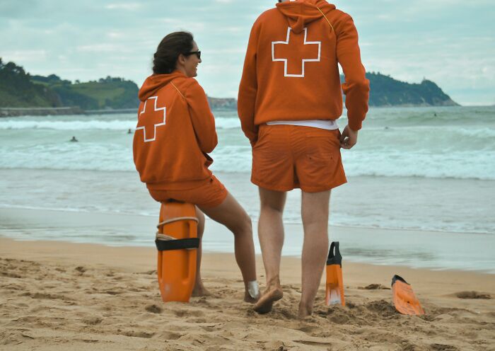 Two travelers, wearing orange hoodies with white crosses, on a beach, experiencing culture shock.