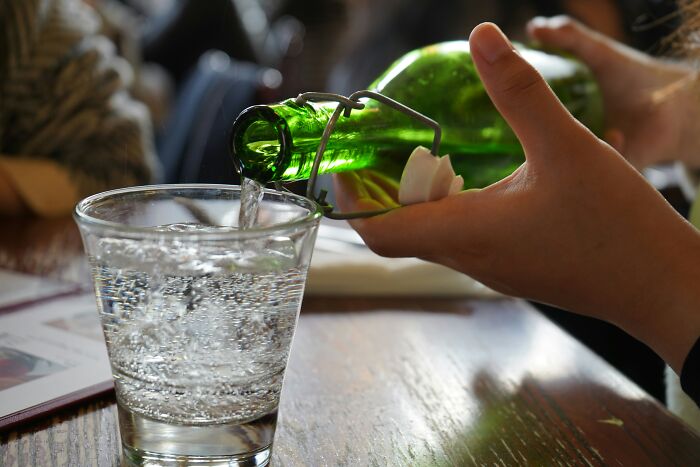 Travelers encountering culture shock from local customs, with a hand pouring sparkling water into a glass from a green bottle.