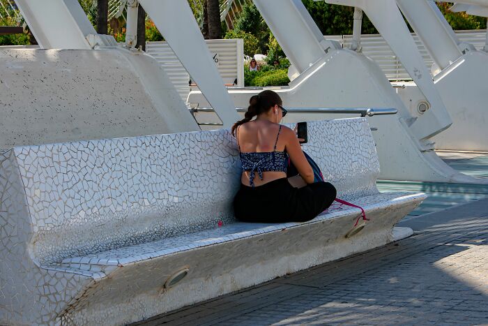 A lone traveler sitting on a modern tiled bench, looking at her phone, possibly experiencing culture shock.