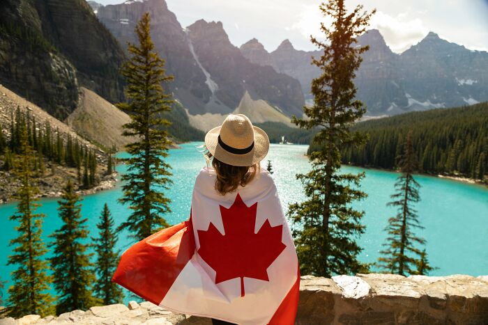 Travelers showing patriotism with a Canadian flag draped over her shoulders, looking at a beautiful lake and mountains.