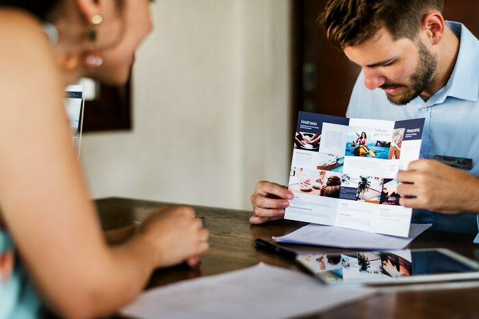 Man showing a brochure with travel packages to a woman, revealing behind the scenes secrets of travel companies.