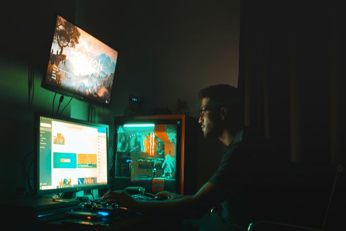 A person concentrating on playing video games on multiple monitors in a dim room, perfect for forgetting facts.