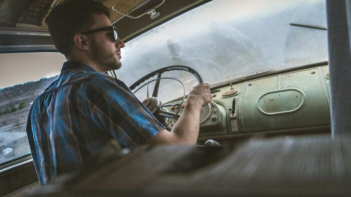 A man driving an old car, his expression suggesting a terrifying 'We Need To Leave Now' situation unfolding on the road.