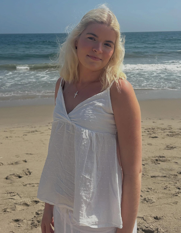 Daughter of former highest-paid TV actor, wearing a white top, stands on a sandy beach by the ocean. Daughter of former highest-paid TV actor, wearing a white top, stands on a sandy beach by the ocean.