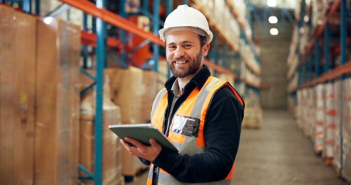 A smiling man in a hard hat and safety vest holding a tablet in a warehouse, showcasing industries that make millions.