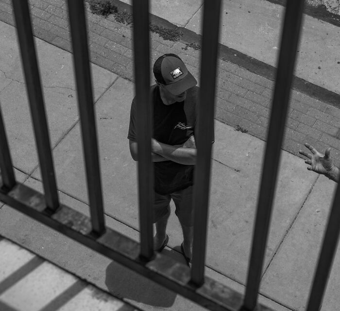 A man viewed through jail bars, his posture hinting at a terrifying 'We Need To Leave Now' moment of confinement.