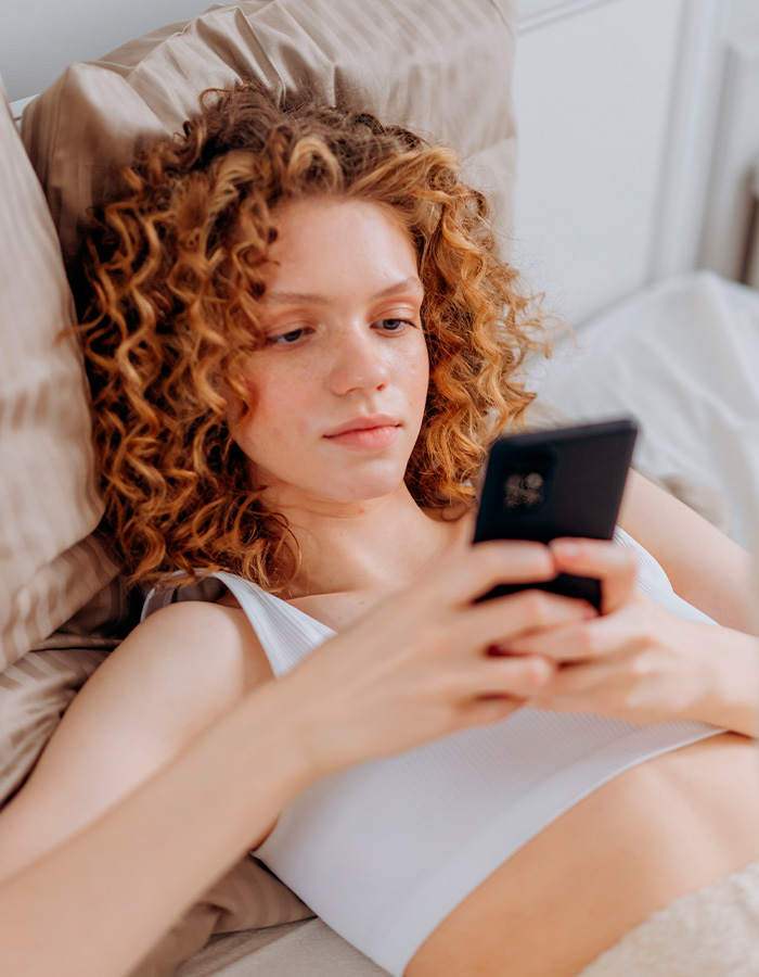 A young woman in bed, engrossed in her phone, highlighting the impact of the new dating method on California colleges. A young woman in bed, engrossed in her phone, highlighting the impact of the new dating method on California colleges.