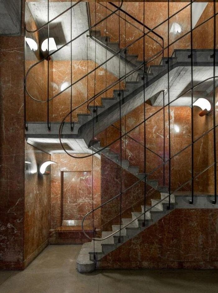 A grand historic interior design featuring a marble staircase with intricate metal railings and wall sconces.