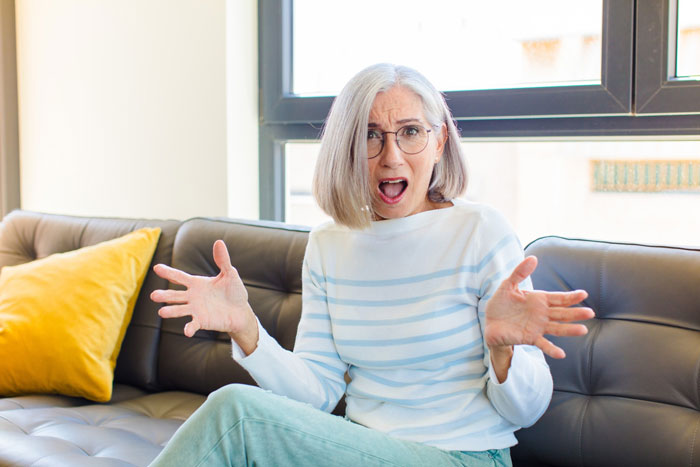 An Entitled Mom looking shocked and upset, sitting on a couch with her hands raised in exasperation. An Entitled Mom looking shocked and upset, sitting on a couch with her hands raised in exasperation.
