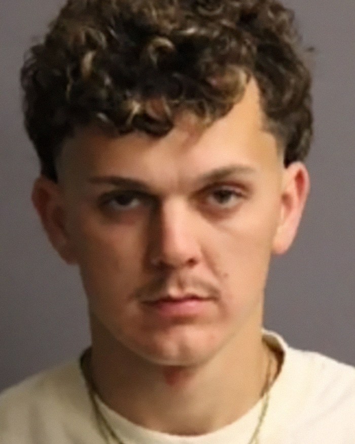 A mugshot of a young man with curly brown hair, light mustache, and a serious expression, related to stupid arrests.
