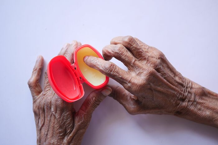 Elderly hands applying ointment from a small container, an everyday item used for different things.