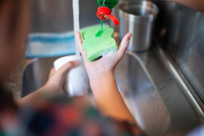 Child pouring dish soap on a sponge, an everyday item used for different things beyond just dishes.