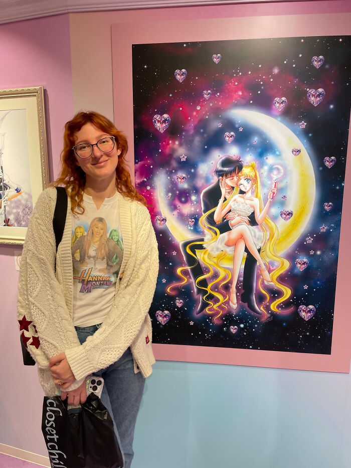 A young woman with red hair standing next to a colorful anime poster, whose looks don't match surprising facts.
