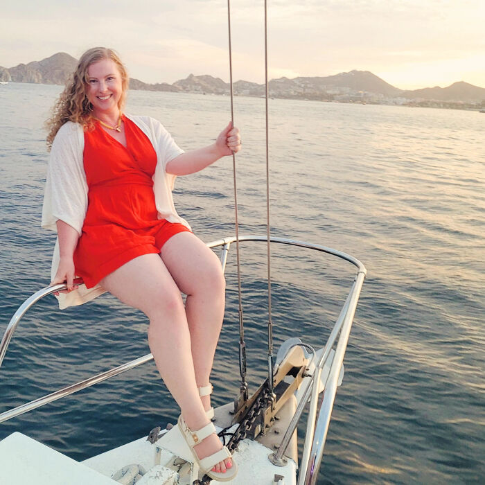 A woman with curly hair in a red romper sitting on the bow of a boat, smiling, revealing surprising facts.