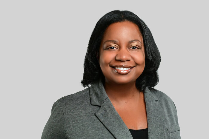 Woman with a broad smile in a grey blazer, black top against a grey background, displaying surprising facts.