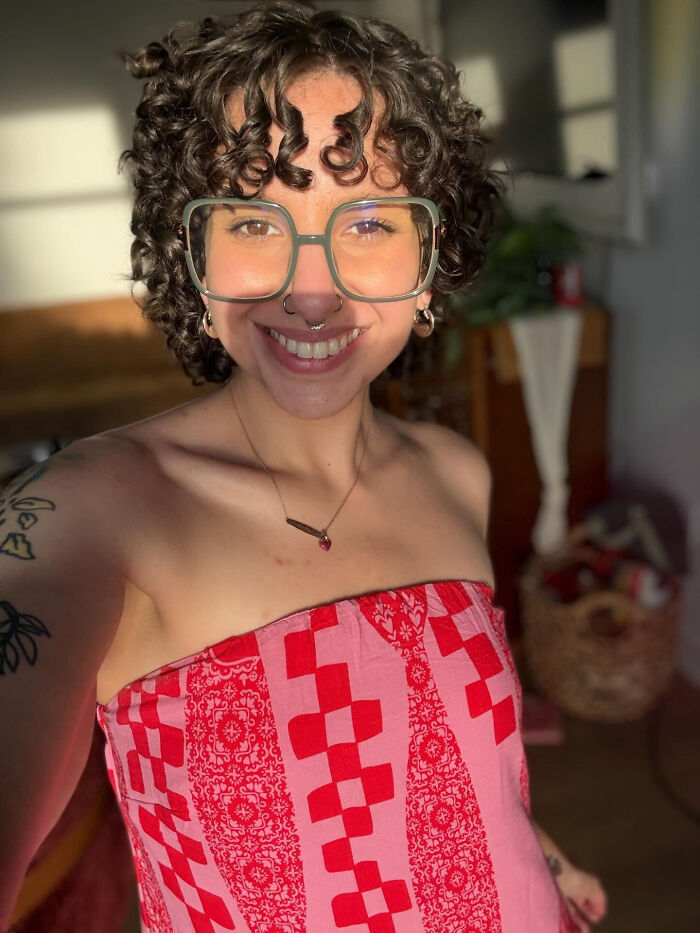 Young woman with curly hair and large glasses, wearing a red and pink patterned top, revealing surprising facts.