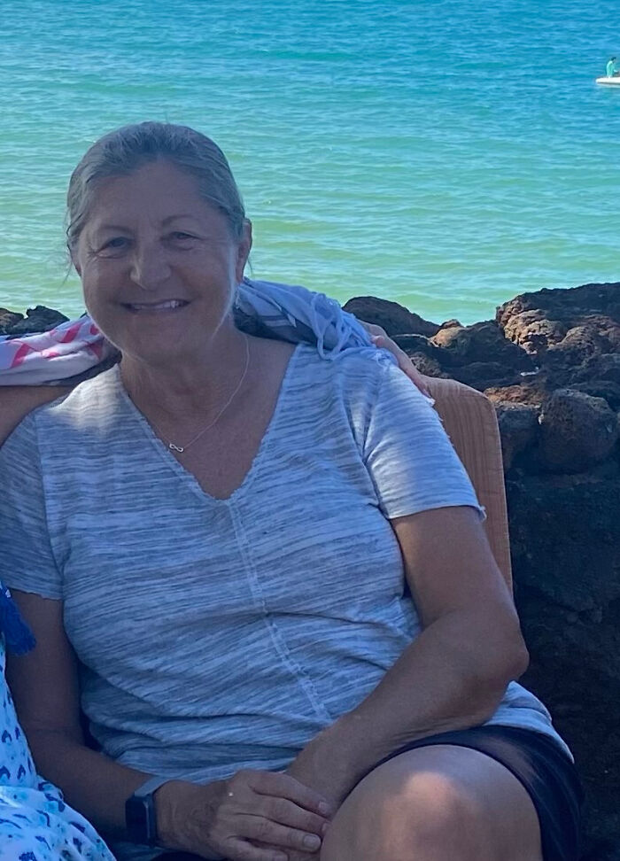 A woman with gray hair, smiling, sitting on rocks by a calm blue ocean. This image contains surprising facts.