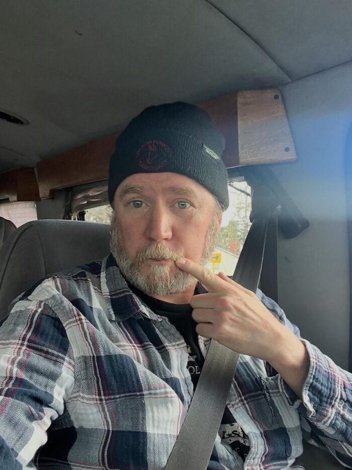 A bearded man in a plaid shirt and black beanie, pointing at his mustache in a car. This image contains surprising facts.