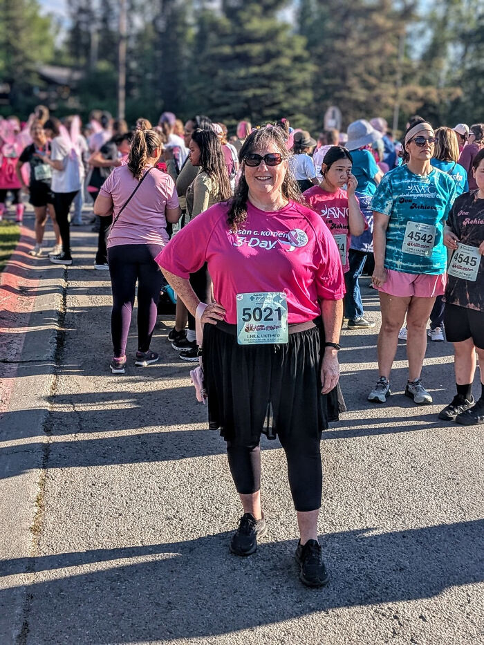 A woman in a pink shirt and black skirt with a race bib, standing on a road during a crowded event. This image contains surprising facts.