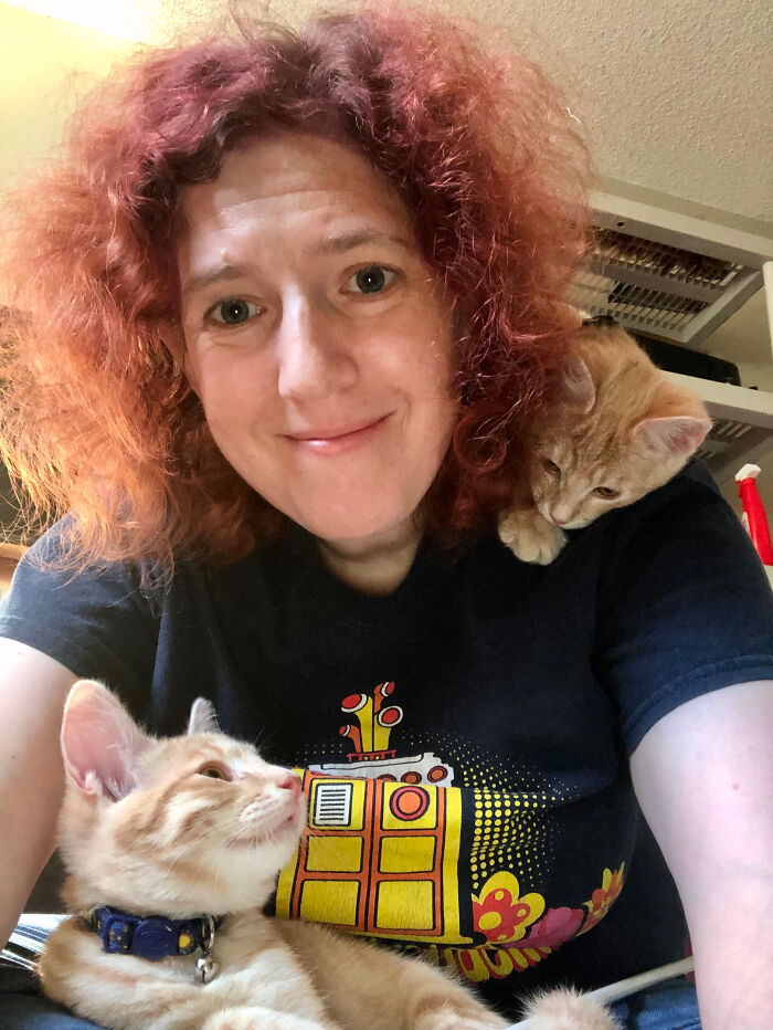 A woman with red, curly hair and a dark t-shirt, smiling at the camera with two ginger kittens. This image contains surprising facts.