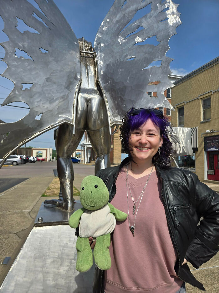 A woman with purple hair holding a turtle plushie next to a metal sculpture. Surprising facts about people's looks.