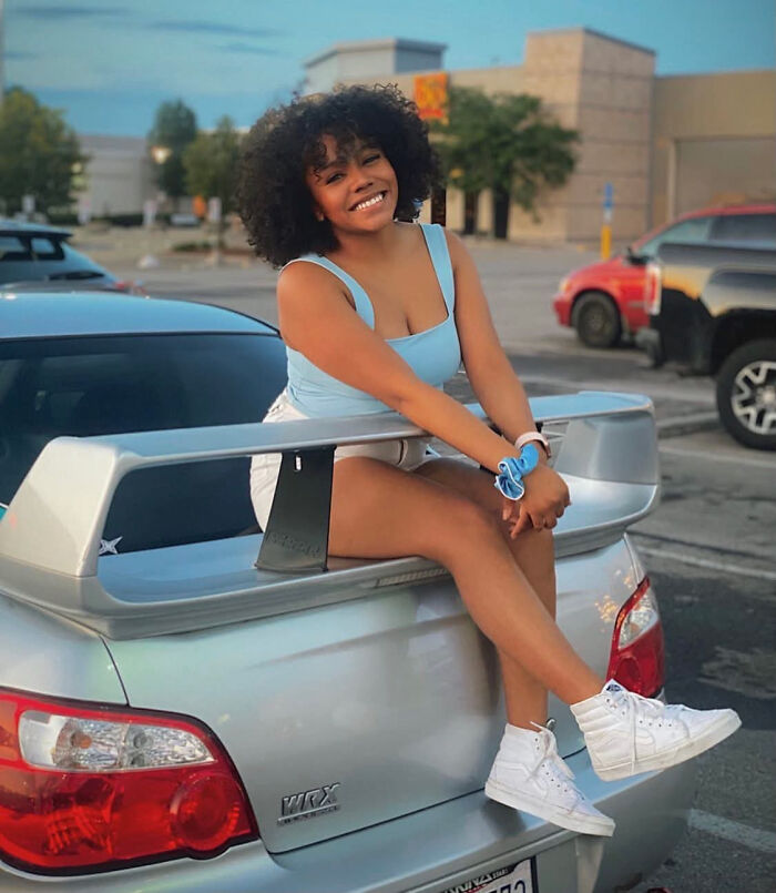 A smiling woman with curly dark hair sitting on a car trunk, representing surprising facts that don't match looks.