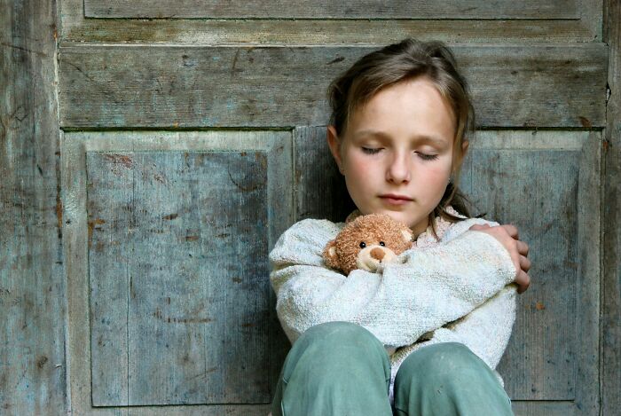 A daughter hugging a teddy bear, eyes closed, against a wooden wall. Her dad's understanding of women evolved.