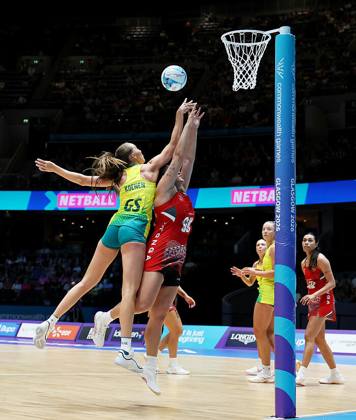 Female athletes in a netball match, one in yellow and one in red, jumping to block a shot near the net.
