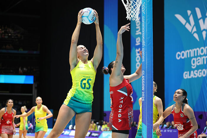 Female athletes in netball match, one in yellow and one in red, reaching for the ball near the hoop.