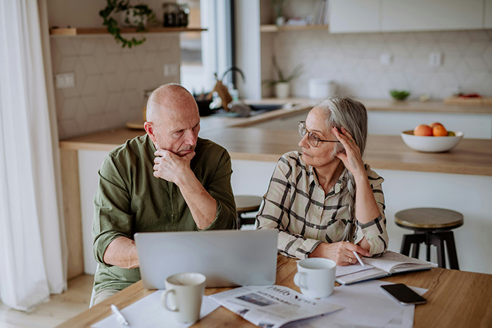An older man and woman at a table with a laptop and papers, discussing financially supporting son in his 40s. An older man and woman at a table with a laptop and papers, discussing financially supporting son in his 40s.