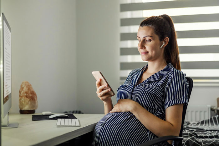 A pregnant woman smiles while looking at her phone, at home, in an image related to a baby name announcement. A pregnant woman smiles while looking at her phone, at home, in an image related to a baby name announcement.