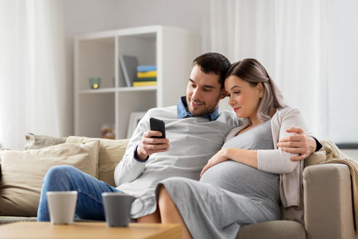 Pregnant woman and man on couch, looking at phone, discussing baby name announcement Pregnant woman and man on couch, looking at phone, discussing baby name announcement