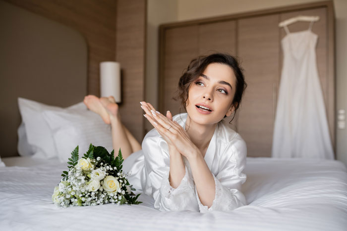 A bride in a white robe on a bed with a wedding dress and bouquet, looking inspired about her violin dream.