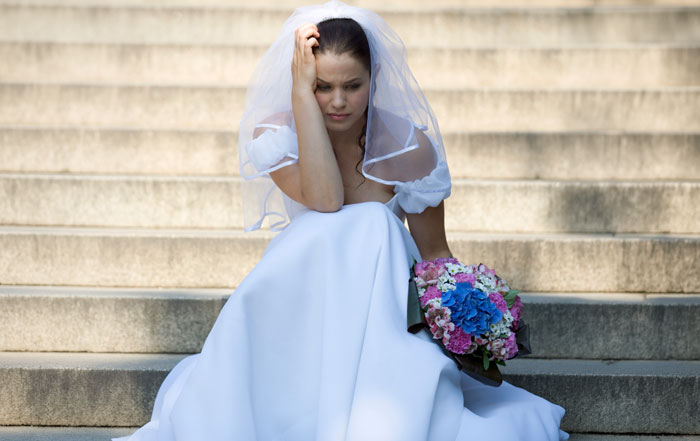A distraught bride in her wedding dress sits on stone steps, head in hand, holding a bouquet after a violin wedding issue.