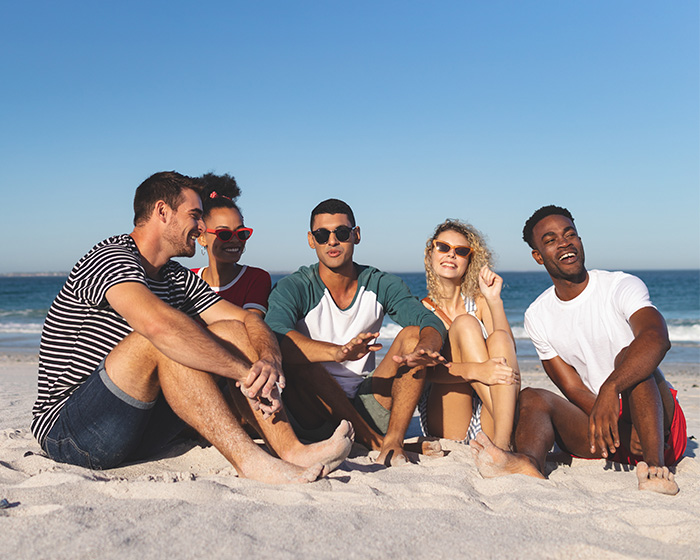 A diverse group of five friends laughing and talking on a sandy beach under a clear blue sky. A diverse group of five friends laughing and talking on a sandy beach under a clear blue sky.