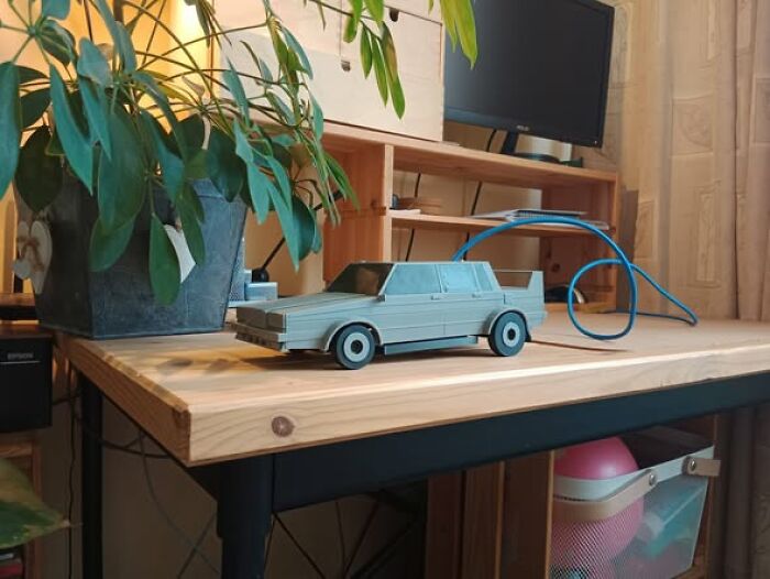 A cool 3D printer creation: a model car on a wooden desk, next to a plant and computer monitor.