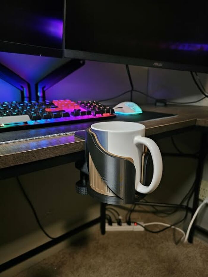 A 3D printer created desk-mounted cup holder with a white mug on a desk with a computer keyboard. It is functional and cool.