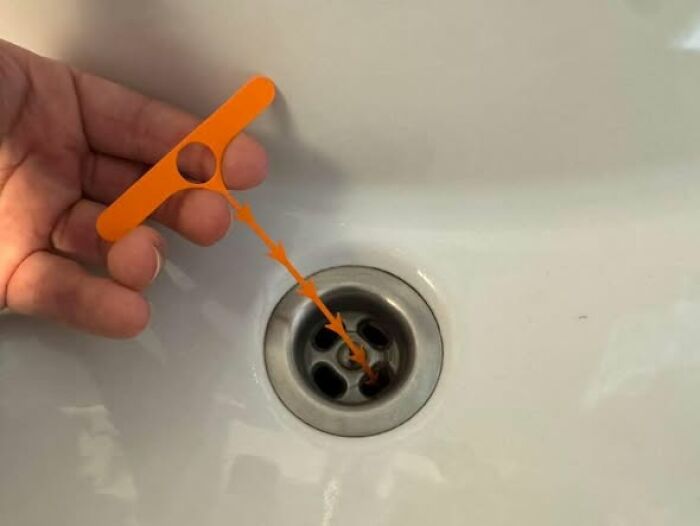 A hand holding an orange drain hair catcher, a functional thing created using a 3D printer, above a sink.