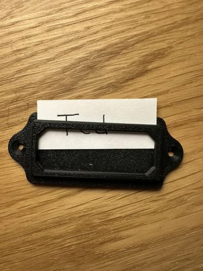 A close-up of a black 3D printed label holder with a paper tag, demonstrating functional 3D printer applications.